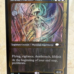 May include: A collectible trading card titled Atraxa, Praetors' Voice, featuring a gold-bordered frame and a central illustration of a multi-winged figure. The card text includes abilities like flying, vigilance, deathtouch, and lifelink.