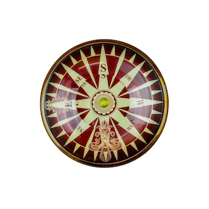 Puede incluir: Una brújula redonda con una esfera roja y crema. La rosa de los vientos presenta marcas direccionales y un diseño central. Está encerrada en un marco de metal dorado. El diseño recuerda a los instrumentos náuticos antiguos.