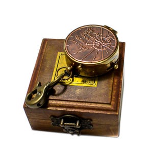 Pode incluir: Uma bússola de estilo vintage com a inscrição "Not All Those Who Wander Are Lost" está em cima de uma caixa de madeira. A bússola é de cor bronze com uma borda dourada e um porta-chaves. A caixa tem uma placa de latão que diz "Sailor's Art".