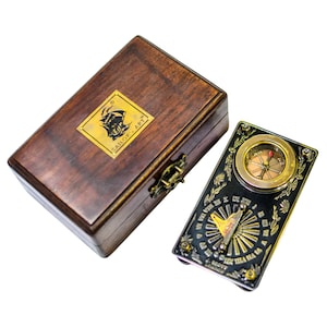 Puede incluir: Un juego de brújula y reloj de sol de estilo vintage en una caja de madera. La brújula tiene un bisel dorado y una esfera negra con detalles ornamentados. La caja es de color marrón oscuro con un cierre de latón y una etiqueta dorada "SAILOR ART".
