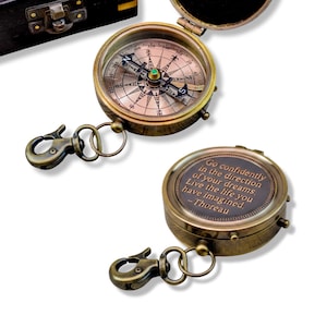 May include: Two antique brass compasses with attached keychains. One compass is open, revealing the directional dial. The other compass has the quote "Go confidently in the direction of your dreams. Live the life you have imagined. - Thoreau" engraved on it.