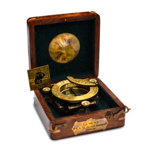 Può includere: Un antico strumento nautico in ottone, probabilmente una meridiana o una bussola, contenuto in una scatola di legno aperta con interno in velluto verde. La scatola presenta una targhetta in ottone con la scritta "J.J. Hicks 12 Hatton Garden London".