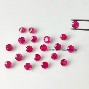 Puede incluir: Una colección de pequeñas piedras preciosas redondas y facetadas de un vibrante color rojo rosado esparcidas sobre una superficie lisa. Una piedra está sujeta por unas pinzas de metal. El texto DwarkaaAllGemstone está impreso en negro en el centro.