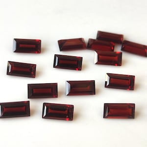 Könnte beinhalten: Eine Sammlung rechteckiger, tiefroter Edelsteine mit polierter Oberfläche. Die Edelsteine sind auf einer weißen Oberfläche verstreut und zeigen ihre satte Farbe und Klarheit. Diese Edelsteine sind ideal für die Schmuckherstellung.