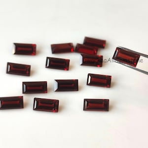 Könnte beinhalten: Eine Sammlung rechteckiger, tiefroter Edelsteine. Ein Edelstein wird von einer Pinzette gehalten. Die Edelsteine sind auf einer weißen Oberfläche verstreut. Die Edelsteine haben eine polierte, reflektierende Oberfläche.