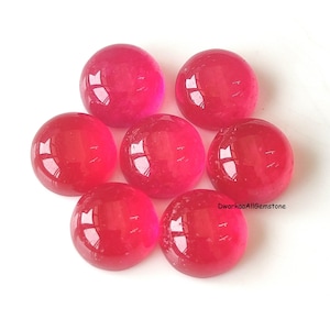 Puede incluir: Siete piedras preciosas redondas, lisas y talladas en cabujón de un color rosa intenso y translúcido. Las piedras están dispuestas en un patrón circular. El texto DwarkaaAllGemstone está impreso en letras negras pequeñas cerca de la piedra inferior derecha.