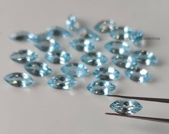 May include: A collection of light blue marquise-cut gemstones. One gemstone is held by tweezers. The gemstones are scattered on a white surface. The gemstones are transparent and have a slight sparkle.