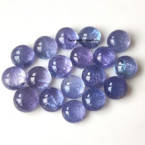 May include: A collection of round, polished gemstones in shades of blue and purple. The stones have a smooth, reflective surface and vary in color intensity. The gemstones are scattered across a white surface.