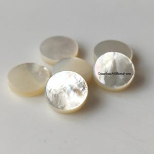 May include: A collection of iridescent, round mother-of-pearl cabochons. The cabochons have a creamy white color with a shimmering, textured surface. The gemstones are arranged on a light-colored surface.
