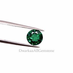 May include: A round, faceted, emerald-green gemstone is held by a pair of silver tweezers against a white background. The gemstone has a brilliant cut, reflecting light. The text "DwarkaaAllGemstone" is visible.