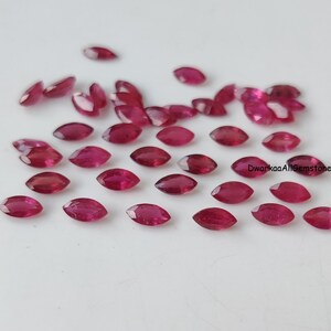 May include: A collection of small, faceted, marquise-cut rubies in a deep red hue. The gemstones are arranged in a loose cluster, displaying a brilliant, reflective surface. The text DwarkaaAllGemstone is printed in black on the right side.
