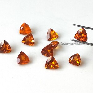 Puede incluir: Colección de gemas triangulares de color naranja. Una gema es sostenida por unas pinzas. Las gemas están dispersas sobre una superficie blanca. Las gemas tienen una superficie pulida y reflectante.