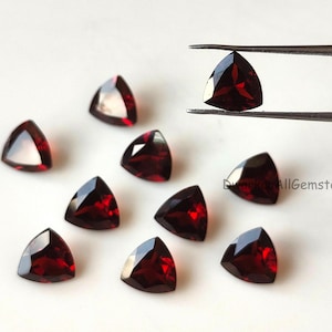 Könnte beinhalten: Eine Sammlung dreieckiger, facettierter, tiefroter Edelsteine. Ein Edelstein wird von einer Pinzette gehalten. Die Edelsteine haben eine polierte, reflektierende Oberfläche. Die Edelsteine sind auf einer weißen Oberfläche angeordnet. Wahrscheinlich Granate.