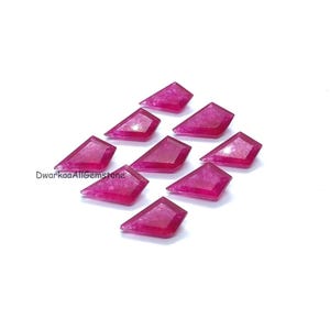 May include: Nine faceted, shield-shaped gemstones in a vibrant magenta hue are arranged on a white surface. The gemstones have a polished, reflective surface, and the edges are sharply defined. The text "DwarkaaAllGemstone" is visible in the lower left corner.