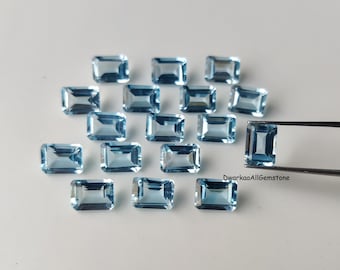 May include: A collection of rectangular, light blue gemstones. One gemstone is held by a pair of tweezers. The gemstones are arranged on a white surface. The text "DwarkaaAllGemstone" is visible.