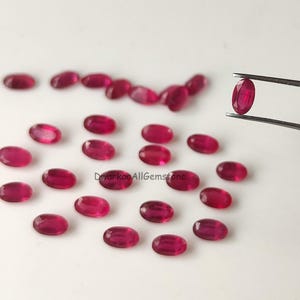 Puede incluir: Una colección de pequeños rubíes de color rojo rosado intenso, con corte ovalado, esparcidos sobre una superficie lisa. Un rubí es sostenido por unas pinzas de metal. El texto DwarkaaAllGemstone está impreso en negro en el centro del conjunto.