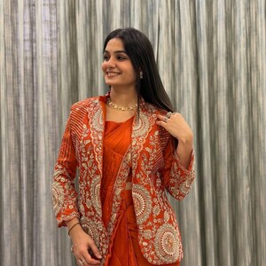 May include: An orange sari and matching jacket with intricate gold embroidery. The jacket features a mandarin collar and long sleeves. The sari is draped elegantly, creating a flowing silhouette. The outfit is accessorized with gold jewelry.