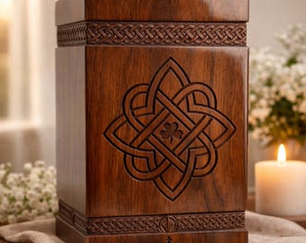 Könnte beinhalten: Dunkelbraune Holzurne mit keltischem Knotenmuster und Kleeblatt. Die rechteckige Urne hat geschnitzte Details und ein geflochtenes Muster oben und unten. Eine brennende Kerze und Blumen im Hintergrund sorgen für eine warme Atmosphäre.