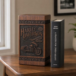 K&ouml;nnte beinhalten: Eine Holzbox mit dem Harley-Davidson-Logo und einem Motorrad-Design mit geschnitzten Details. Daneben ein schwarzes Buch mit dem Titel "A Prayer for Owen Meany" von John Irving.