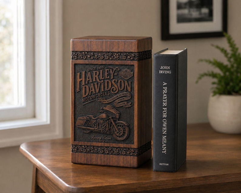 K&ouml;nnte beinhalten: Eine Holzbox mit dem Harley-Davidson-Logo und einem Motorrad-Design mit geschnitzten Details. Daneben ein schwarzes Buch mit dem Titel "A Prayer for Owen Meany" von John Irving.