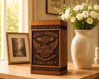 Könnte beinhalten: Eine rechteckige Holzbox mit einem geschnitzten Harley-Davidson-Logo mit Flügeln und dem Schriftzug "SINCE 1903". Die Box hat dekorative florale Ränder oben und unten sowie ein dunkelbraunes, strukturiertes Mittelteil mit dem Emblem.