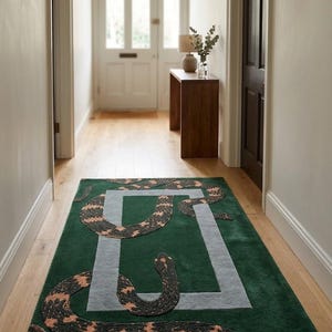 Può includere: Un tappeto rettangolare con uno sfondo verde intenso e un bordo rettangolare grigio chiaro. Il tappeto presenta un grande motivo a serpente sinuoso nei toni del nero e del rame, che si snoda attorno e attraverso la cornice centrale grigia.