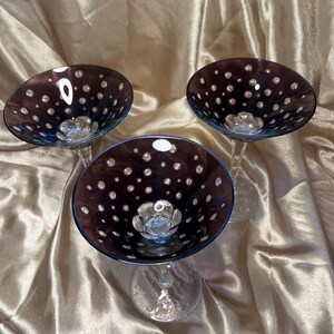 May include: Three stemmed cocktail glasses with a dark purple bowl dotted with white spots. The rim of each glass is a deep blue. The glasses are clear and sit on a gold-colored surface.
