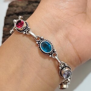 Pode incluir: Uma pulseira de metal prateado com uma série de engastes ovais e redondos. Os elos são decorados com padrões intrincados de fio torcido e contêm pedras preciosas facetadas em tons vibrantes de vermelho, azul brilhante e branco transparente.