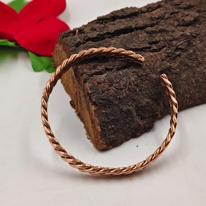 May include: A copper-colored, braided cuff bracelet. The bracelet is open at one end and rests on a piece of dark brown wood. A red flower is visible in the background.