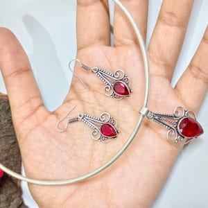 Może przedstawiać: Srebrny zestaw biżuterii składający się z naszyjnika i pasujących kolczyków. Zestaw zawiera naszyjnik typu choker, wisiorek i dwie kolczyki wiszące. Każdy element ma łezkowaty, ciemnoczerwony kamień szlachetny. Biżuteria jest prezentowana na dłoni.