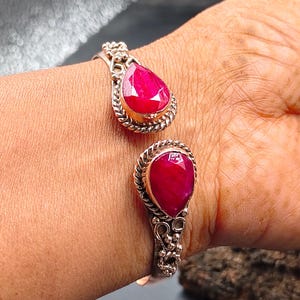 May include: A silver cuff bracelet with two large, teardrop-shaped, ruby-red gemstones. The gemstones are set in a decorative, rope-like border. The bracelet is open and adjustable, with ornate silver detailing.
