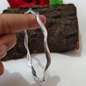 May include: A silver-coloured, wavy bangle bracelet. The bracelet has a fluid, organic design with a polished finish. The bracelet is held up by a person's fingers. The background includes a piece of wood and a red flower.
