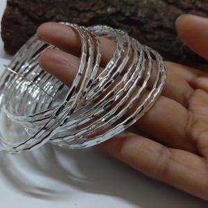Puede incluir: Un conjunto de pulseras plateadas con un diseño texturizado y martillado. Las pulseras están apiladas, reflejando la luz y creando un efecto brillante. Las joyas se sostienen en una mano, mostrando los intrincados detalles de las pulseras.