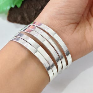 May include: A set of five silver-colored, flat, and wide bangles are stacked on a wrist. The bracelets have a smooth, reflective surface, and are worn on the wrist. The background is blurred, with hints of green and red.