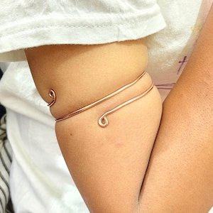 May include: A copper-colored arm cuff with a spiral design. The cuff wraps around the upper arm with two parallel bands, featuring a looped end. The jewelry is worn on a person's arm, against a white shirt.
