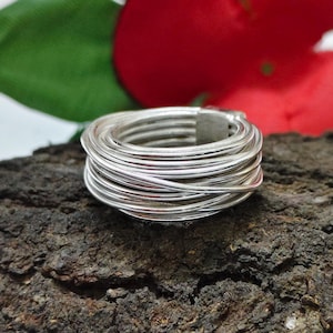 Op de afbeelding: Een zilveren ring met een gevlochten draadontwerp. De ring is gemaakt van meerdere strengen zilverdraad, waardoor een getextureerd en gelaagd uiterlijk ontstaat. De ring is geplaatst tegen een donkere, getextureerde achtergrond.