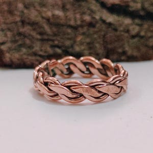Puede incluir: Un anillo de color cobre con un diseño trenzado. El anillo está hecho de hebras de metal entrelazadas, creando una apariencia texturizada y tejida. Se encuentra sobre una superficie blanca con un fondo de madera borroso.