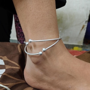 May include: A silver-colored anklet with two curved bands, each adorned with small, round embellishments. The anklet is worn around the ankle, showcasing its simple yet elegant design. The jewelry is a delicate accessory.