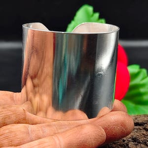 May include: A wide, polished silver cuff bracelet. The bracelet has a smooth, reflective surface and a simple, modern design. The cuff is approximately 5 cm wide.