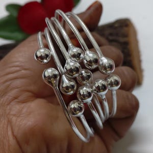 May include: A collection of silver-toned bangles, each featuring a smooth, spherical accent. The bangles are displayed against a neutral background, with a hint of red and brown in the periphery. The jewelry is designed with a minimalist aesthetic, suitable for everyday wear.