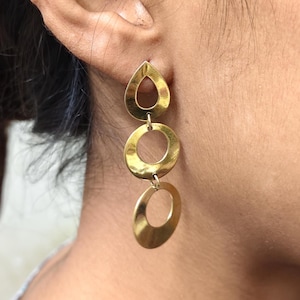 May include: Gold-toned earrings with a teardrop shape at the top, connected to two circular hoops. The earrings are dangling and have a polished finish, reflecting light. The jewelry is worn on an ear.