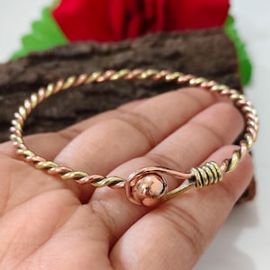Puede incluir: Un brazalete trenzado hecho de hilos alternos de cobre y latón. La pulsera presenta un cierre de estilo gancho con una bola de cobre pulido en el extremo, asegurada por un detalle de alambre de latón enrollado.