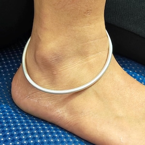 Puede incluir: Una tobillera plateada rodea un tobillo. La tobillera es una banda lisa y sencilla. El tono de piel es claro y el fondo es una superficie azul con un patrón de puntos blancos. La tobillera parece ser una pieza de joyería minimalista.