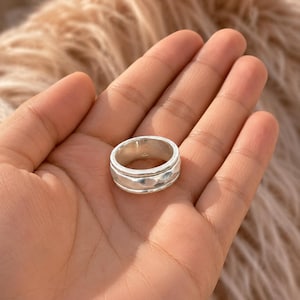 May include: A silver-colored ring with a textured band and a smooth, rotating center. The ring is round and appears to be made of metal. The ring is shown on a hand.