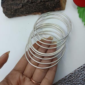 Può includere: Una pila di braccialetti color argento. I braccialetti sono rotondi e sembrano essere di metallo. I gioielli sono impilati l'uno sull'altro. I braccialetti sono tenuti da una persona.