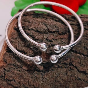 May include: Two silver-colored, open-ended bangles with a smooth, rounded ball at each end. The bangles are displayed on a textured, brown wooden surface. The jewelry is simple and elegant.