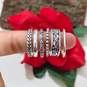 May include: A set of six silver-toned rings featuring various designs, including a braided pattern, a smooth band, a geometric etched band, a beaded band, a wider band with a floral motif, and a plain thin band, all displayed in a row.