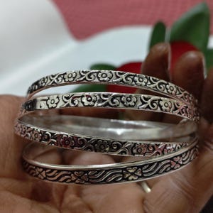 Può includere: Un set di braccialetti color argento con intricati disegni floreali e a volute. I braccialetti sono impilati, mostrando i modelli dettagliati. I gioielli sono tenuti in una mano, con uno sfondo sfocato rosso e verde.