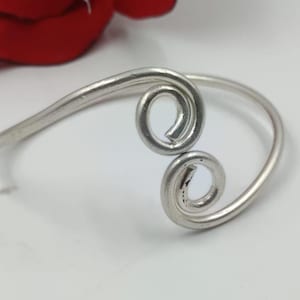 May include: A silver cuff bracelet with a distinctive design. The bracelet has two spiral features and a curved band. The jewellery is displayed on a white surface with a red flower in the background.