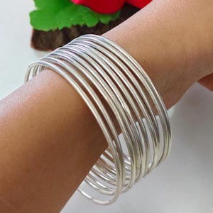 May include: A collection of silver-colored bangles stacked on a wrist. The bangles are thin and round, reflecting light. The jewelry is displayed against a soft background, with a glimpse of a red flower and green leaves.